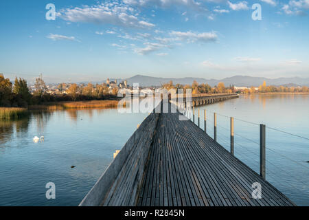 landscape view of marsh and lake shore with the town of Rapperswil in evening light and a long wooden boardwalk in the foreground and a couple going f Stock Photo