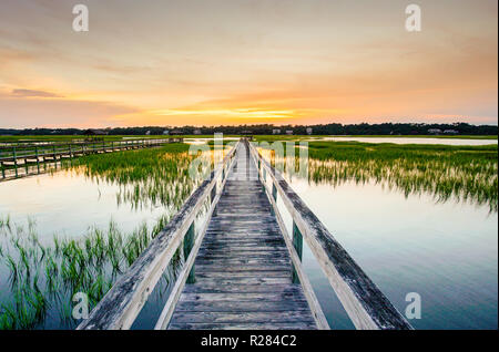 coastal waters with a very long wooden boardwalk pier in the center during a colorful summer sunset under an expressive sky with reflections in waterP Stock Photo