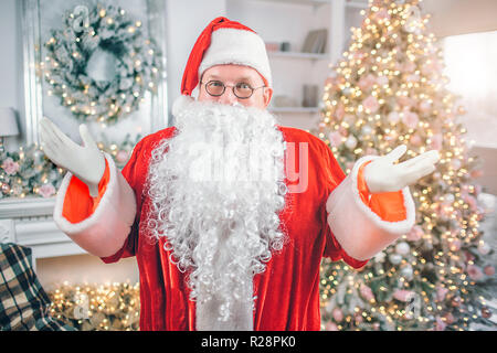 Happy Santa Claus stands and looks on camea. He poses with hands aside of body. Man smiles. There are festive Christmas tree and fireplace behind him. Stock Photo