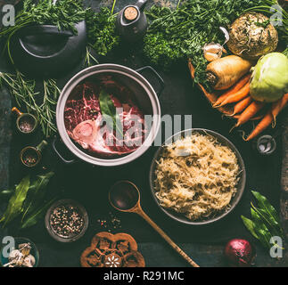 Raw beef meat shin with bone in cooking pot with spices and bowl with pickled cabbage on dark kitchen tables background with low carb ingredients, top Stock Photo