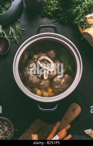 Broth of beef meat shin with bone in cooking pot on dark kitchen table background with ingredients for soup, top view. Meat broth or stock. Clean low- Stock Photo