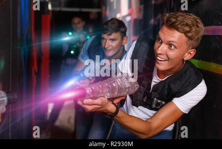 Young positive cheerful glad smiling guy holding colored laser guns and took aim during laser tag game in labyrinth Stock Photo