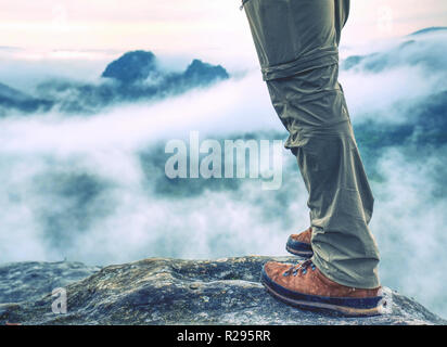 Hiker legs in light outdoor trousers and comfortable leather  trekking boots on rocky peak. Misty valley below peak in far blurry background. Hiking i Stock Photo