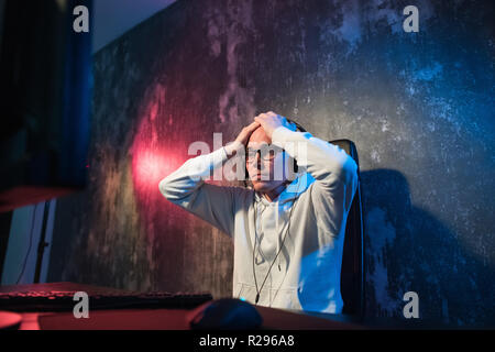 Funny young gamer with a headset playing video games on a computer and looks at the camera with astonishment. Portrait of a surprised young man using  Stock Photo