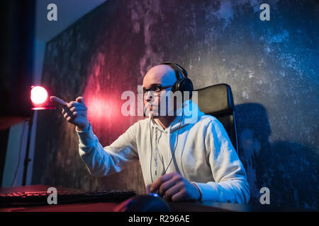 Funny young gamer with a headset playing video games on a computer and looks at the camera with astonishment. Portrait of a surprised young man using  Stock Photo