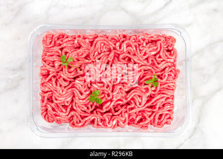 A photo of minced meat in a plastic tray, shot from above on a white marble background with parsley leaves Stock Photo