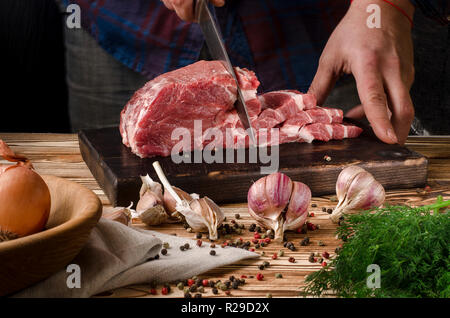 Man slicing pork on wooden board at restaurant kitchen. Chef preparing fresh meat for cooking. Modern cuisine backgroung with herbs and vegetables veg Stock Photo