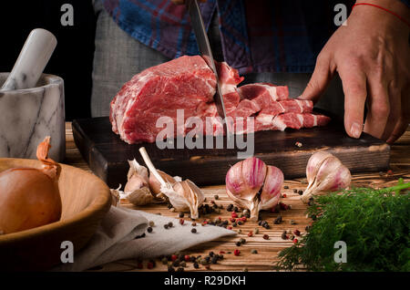 Man slicing pork on wooden board at restaurant kitchen. Chef preparing fresh meat for cooking. Modern cuisine backgroung with herbs and vegetables veg Stock Photo