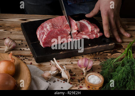 Man slicing pork on wooden board at restaurant kitchen. Chef preparing fresh meat for cooking. Modern cuisine backgroung with vegetables Stock Photo