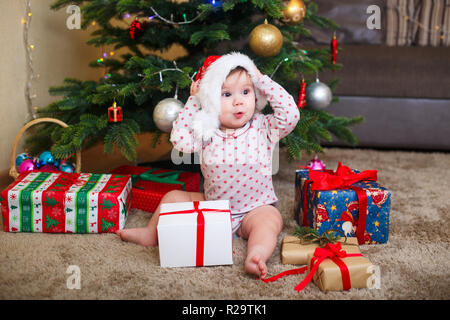 Happy cute baby girl in Santa Claus hat holding Christmas gifts at christmas tree at home Stock Photo