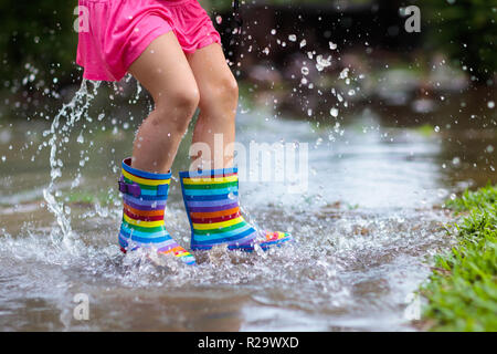 Kid playing out in the rain. Children with umbrella and rain boots play outdoors in heavy rain. Little girl jumping in muddy puddle. Kids fun by rainy Stock Photo