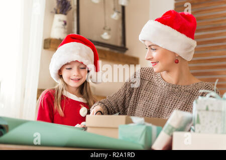 Mother and daughter wearing santa hats having fun wrapping christmas gifts together in living room. Candid family christmas time lifestyle background. Stock Photo