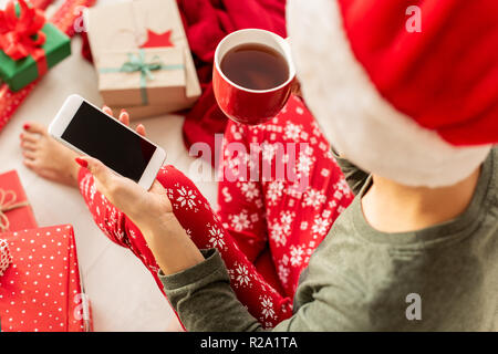Young woman wearing santa hat and xmas pajamas sitting on the floor amongst wrapped christmas presents, drinking hot punch and shopping online on her  Stock Photo
