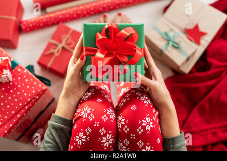Young woman wearing xmas pajamas sitting on the floor amongst wrapped christmas presents, holding beautiful gift box with large red ribbon bow. Person Stock Photo