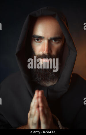 Medieval monk in black robe with hood praying with closed eyes, secret ritual. Mystery and spirituality Stock Photo