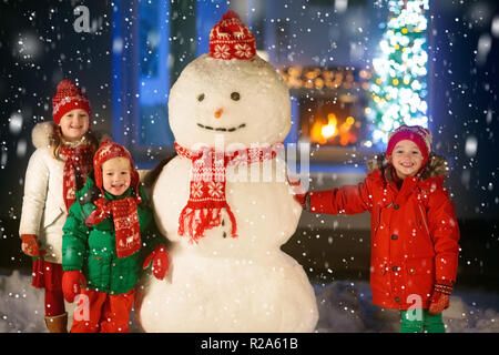 Kids build snowman on Christmas eve. Children in snowy backyard next to window to living room with Christmas tree and decorated fireplace. Boy and gir Stock Photo