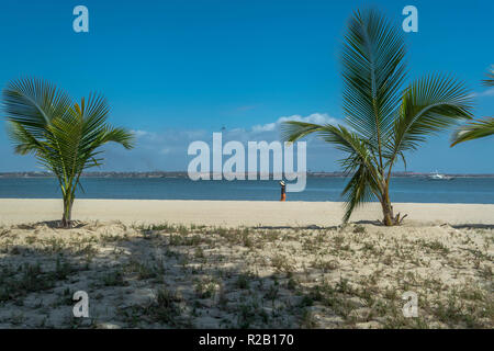 View of palm trees and a woman on beach, and boats on water, on the island of Mussulo, Luanda, Angola... Stock Photo