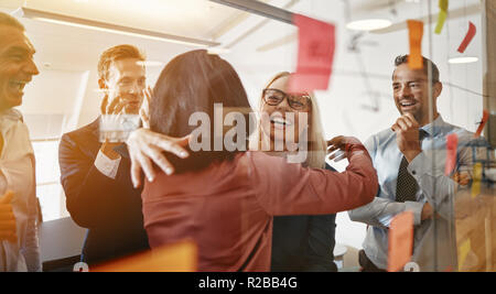 Two smiling young businesswomen hugging while brainstorming with sticky notes on a glass wall with colleagues in a modern office Stock Photo