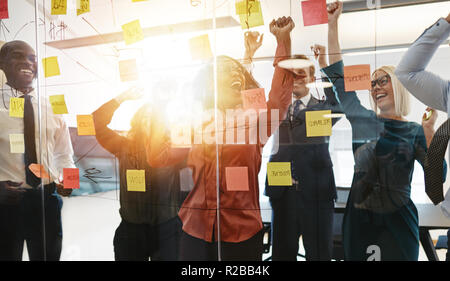 Ecstatic group of diverse businesspeople yelling and jumping up and down while brainstorming with sticky notes on a glass wall in a modern office Stock Photo