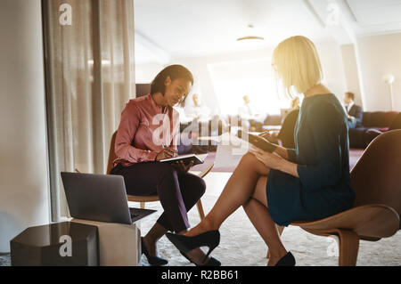 Two smiling female work colleagues sitting together in a bright modern office discussing notes and using a laptop Stock Photo