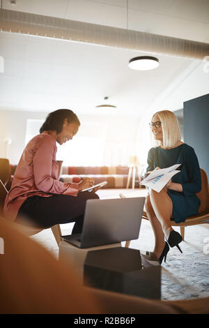 Two young businesswomen smiling while going over documents and working on a laptop together in a modern office Stock Photo