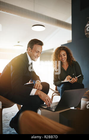 Two diverse young businesspeople smiling while stting in an office talking and working online together with a laptop Stock Photo