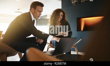Two diverse young businesspeople sitting together in a modern office discussing work over a laptop Stock Photo