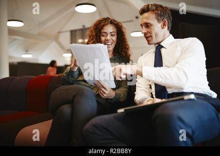 Two diverse work colleagues sitting on a sofa in a modern office discussing a project together Stock Photo