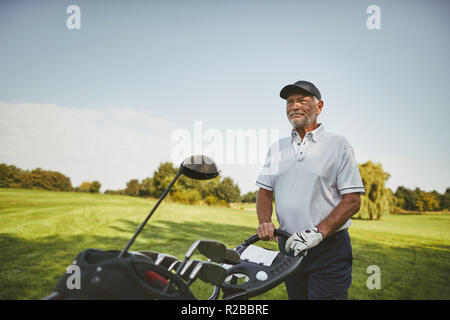 Smiling senior man walking along a fairway with his club bag while playing a round of golf on a sunny day Stock Photo