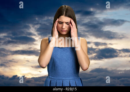 Caucasian woman with terrible migraine. Beautiful upset girl suffering from headache on evening sky background. Human expressions of pain. Stock Photo