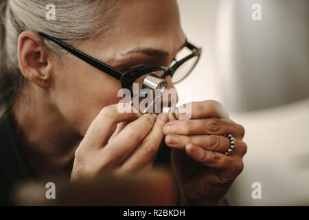 Mature female jeweler looking at stone through loupe to inspect it. Close up of senior jewelry maker looking at the gem while working at workshop. Stock Photo