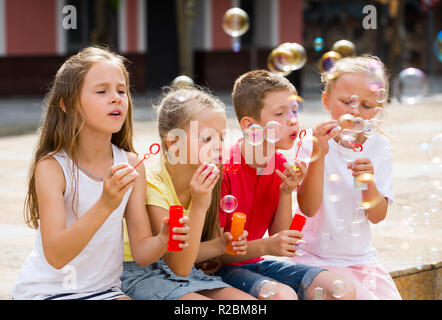 Four children sit in playground and blowing soap bubbles Stock Photo