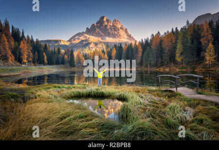 Man with raised up arms on Antorno lake with reflection of Tre Cime Di Lavaredo at sunrise in autumn in Dolomites, Italy. Landscape with happy man, gr Stock Photo