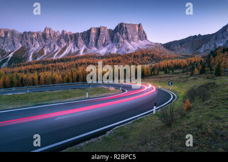 Blurred car headlights on winding asphalt road in mountains at sunset in autumn. Spectacular landscape with light trails, forest, rocks and purple sky Stock Photo