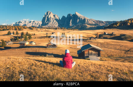 Sitting woman on the hill looking on meadows and mountains at sunset in autumn. Alpe di Siusi, Dolomites, Italy. Landscape with girl in red jacket, fi Stock Photo