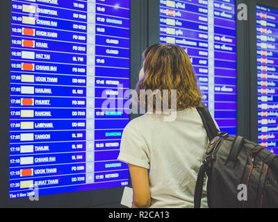 Woman short hair wearing white shirt with backpack checking flight at the flight information board in international airport terminal. Stock Photo