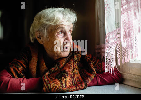 Elderly woman sitting in the house looks longingly out the window. Stock Photo
