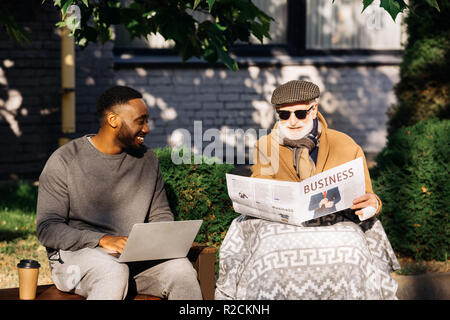 happy senior disabled man reading business newspaper in wheelchair while african american man using laptop on street Stock Photo