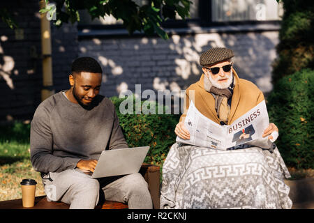 senior disabled man reading business newspaper in wheelchair while african american man using laptop on street Stock Photo