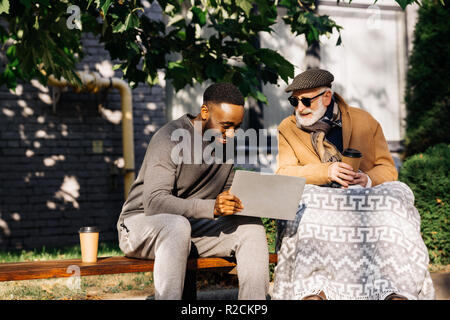 senior disabled man in wheelchair and african american man using laptop together on street Stock Photo