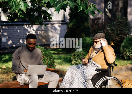 senior disabled man in wheelchair and young african american man using gadgets together on street Stock Photo
