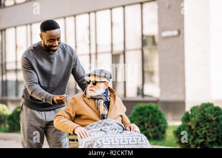 happy african american man giving joint to senior disabled man in wheelchair wjhile they spending time together on street Stock Photo
