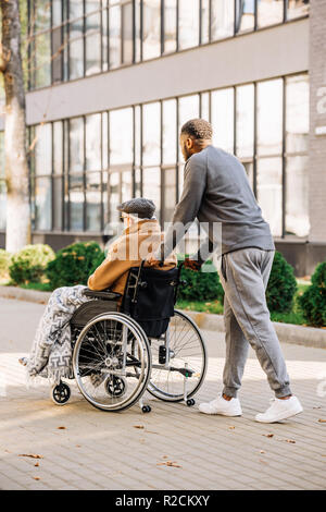 senior disabled man in wheelchair with plaid and cuidador riding by street Stock Photo