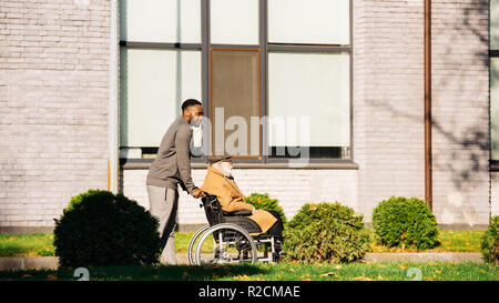 senior disabled man in wheelchair and african american man having morning walk on street Stock Photo
