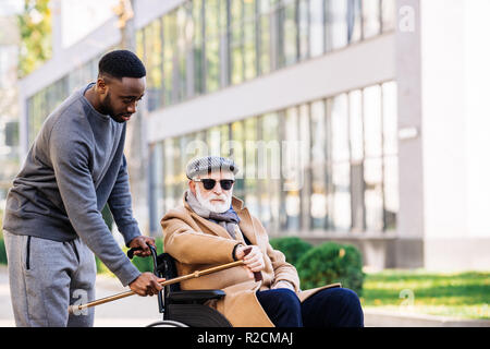 african american man giving walking stick to senior disabled man in wheelchair on street Stock Photo