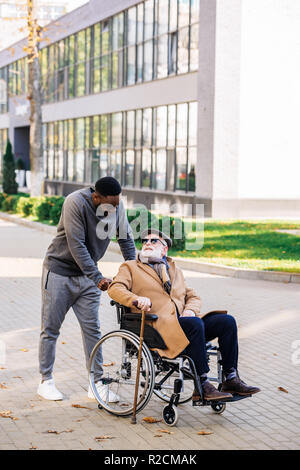 senior disabled man in wheelchair with walking stick and african american cuidador talking on street Stock Photo