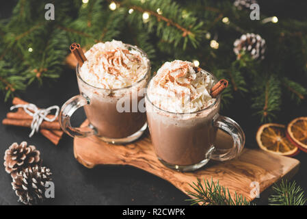 Two cups of hot chocolate with whipped cream and cinnamon on wooden serving board surrounded with fir tree, Christmas lights, cinnamon and pine cones. Stock Photo
