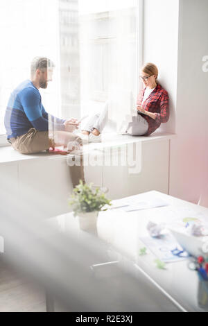Hipster employees using gadgets while sitting on window sill Stock Photo