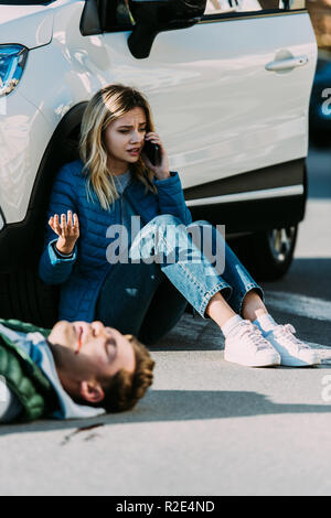 scared young woman calling emergency while injured man lying on road after traffic accident Stock Photo