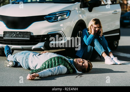 scared young woman calling emergency while injured man lying on road after traffic collision Stock Photo